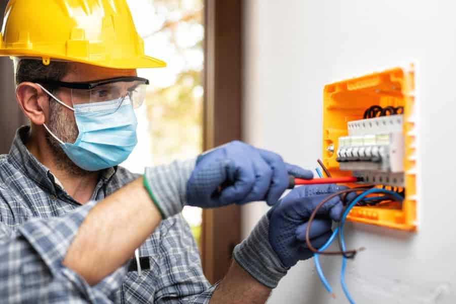 Masked Electrician With Hardhat Working On Electrical Wall Panel